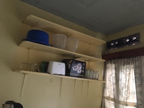 Ein Zimmer mit beige farbenen Wänden, einer Decke mit Gittermuster und einem Fenster mit floralen Vorhängen. Es gibt Regale mit verschiedenen Gegenständen, darunter ein blauer Hut, Plastikbehälter und ein weißes Gerät.