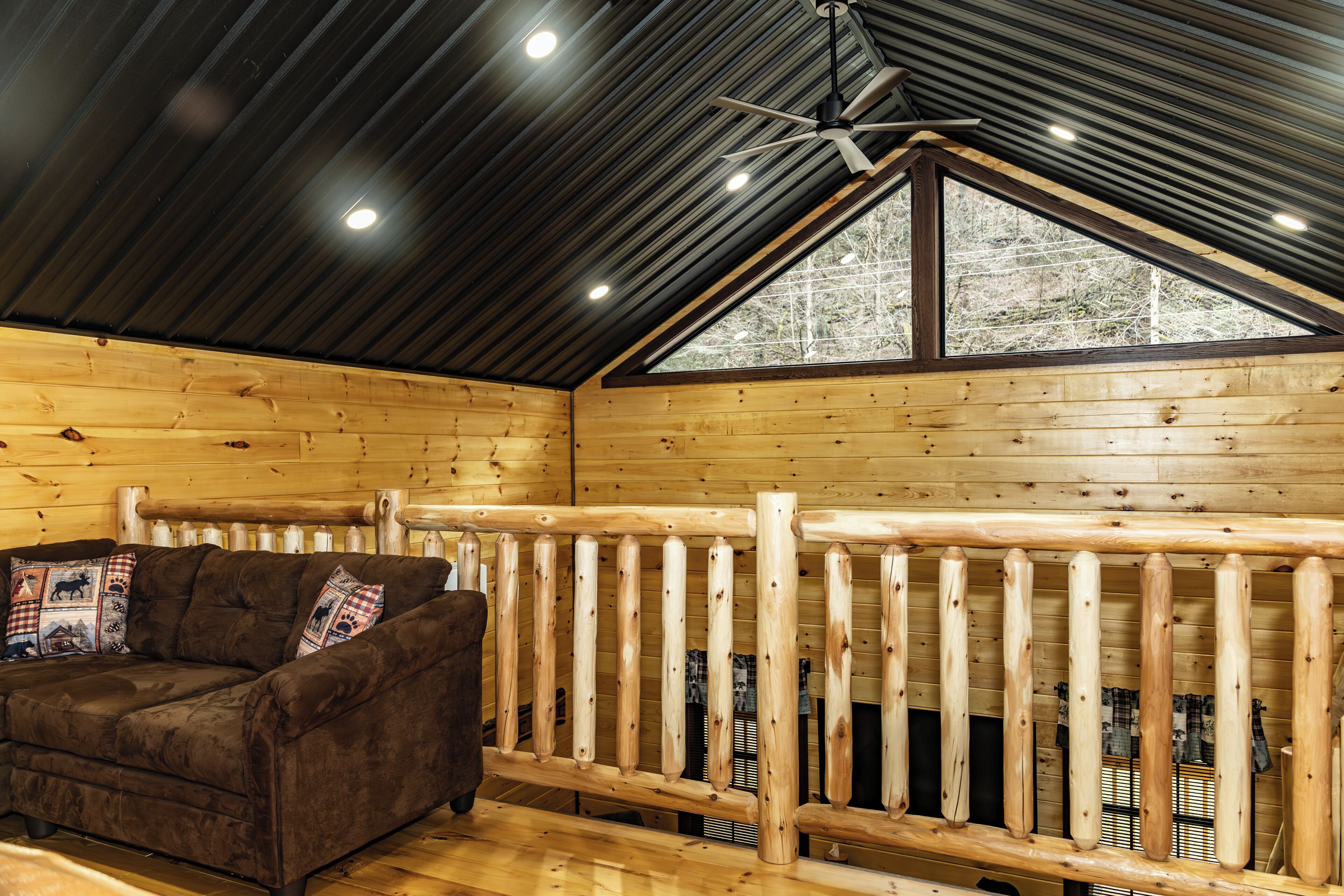 A log cabin interior with a wooden ceiling, a ceiling fan, a window, and a brown sofa with pillows.
