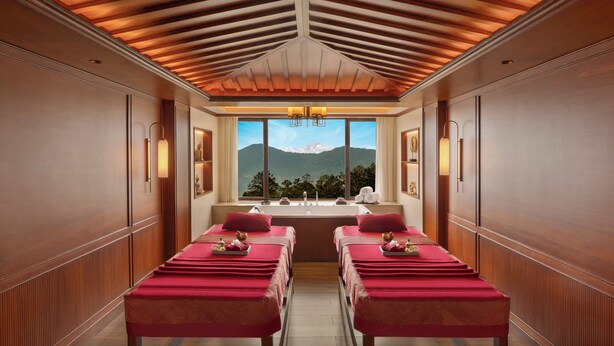 A spa room with wooden paneling, two massage tables, a large window with a mountain view, and wall-mounted lighting. - Welcomhotel By ITC Hotel Denzong Regency (Gangtok)