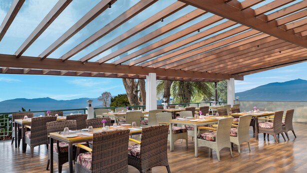 An outdoor dining area with wooden tables and wicker chairs, overlooking a mountainous landscape. - Welcomhotel By ITC Hotel Denzong Regency (Gangtok)