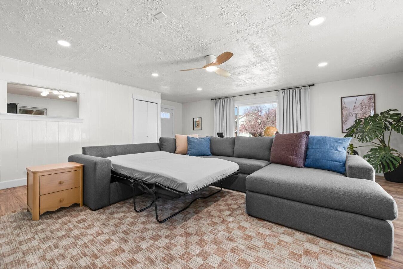 Ein modernes Wohnzimmer mit einem Ecksofa, einem kleinen Nachttisch, einem Deckenventilator und einem großen Fenster mit Vorhängen.