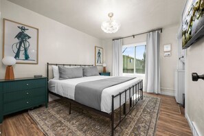 A bedroom with a bed, nightstands, a chandelier, a window with curtains, and artwork on the walls.