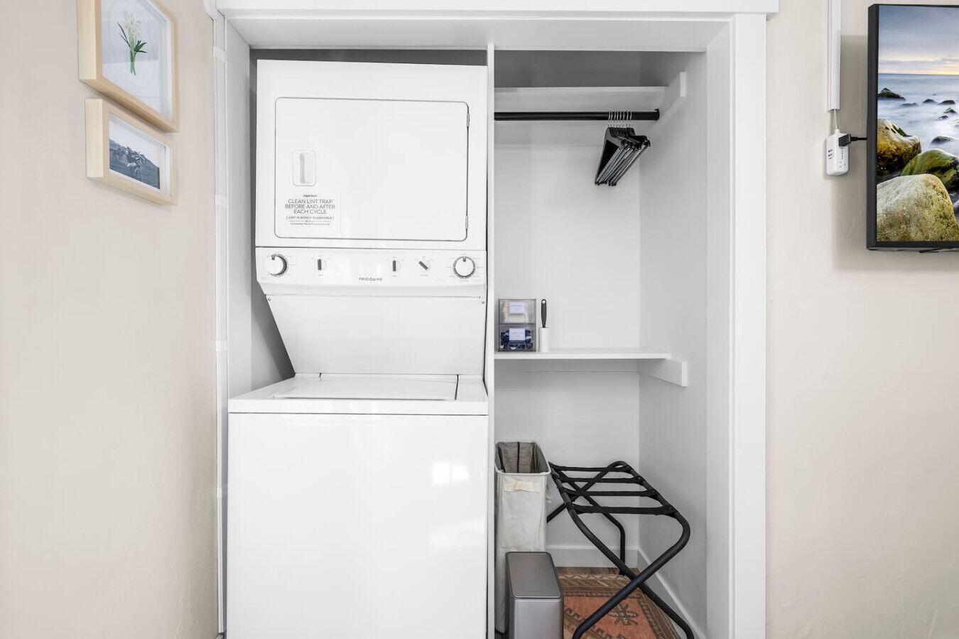 A compact laundry room with a front-loading washer and dryer, a small shelf with a bottle and a knife, and a wall-mounted TV displaying a coastal scene.