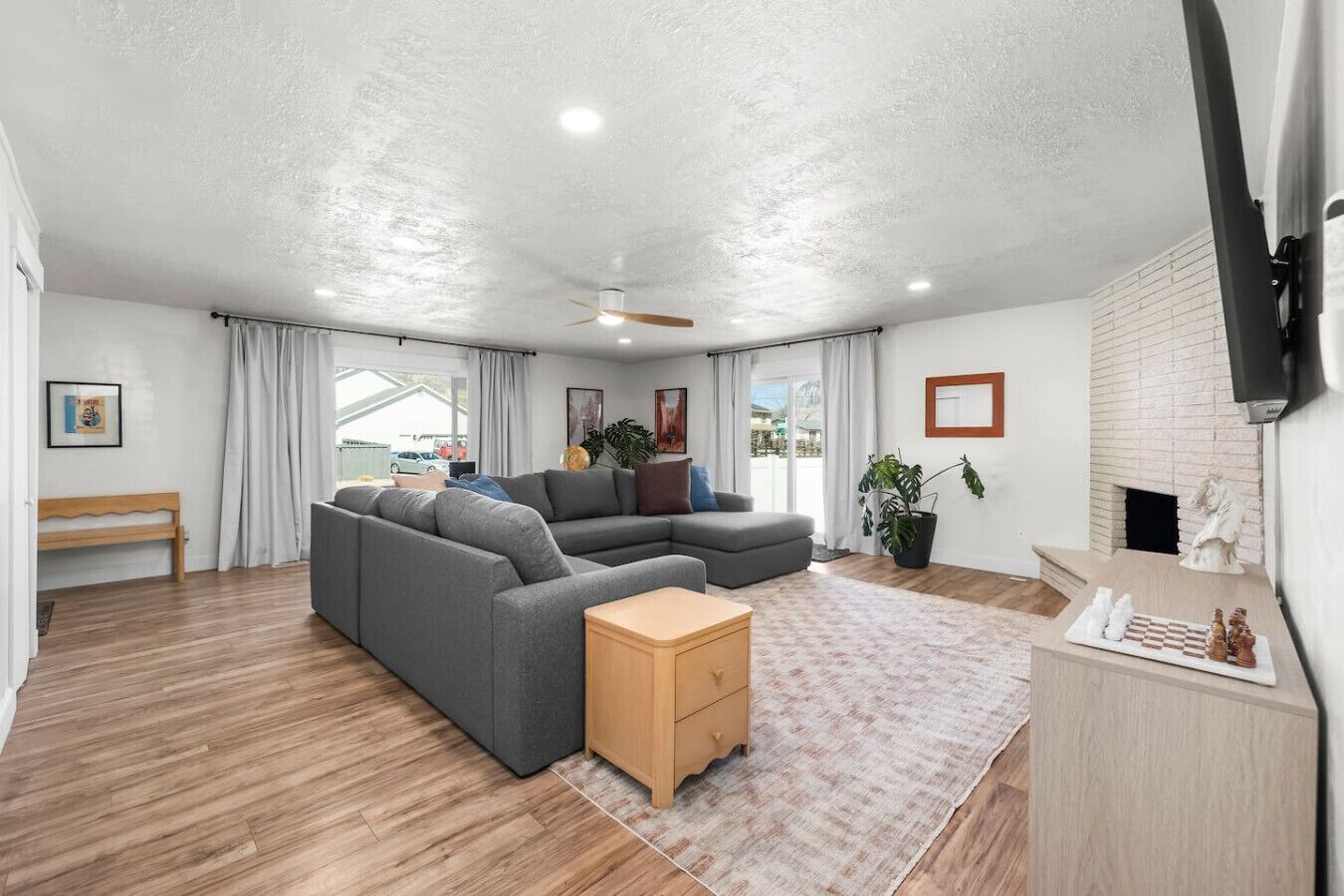 A modern living room with a sectional sofa, a small wooden side table, a fireplace, and a large window with curtains.