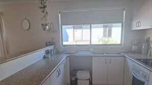 Una cocina con armarios blancos, encimera de granito, ventana con persianas y un pequeño cubo de basura.