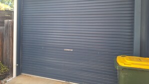Puerta de garaje cerrada con un bote de basura verde a su lado.