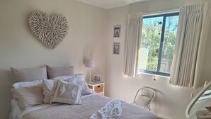 Un dormitorio con cama, decoración en forma de corazón en la pared, mesita de noche con lámpara y ventana con cortinas.