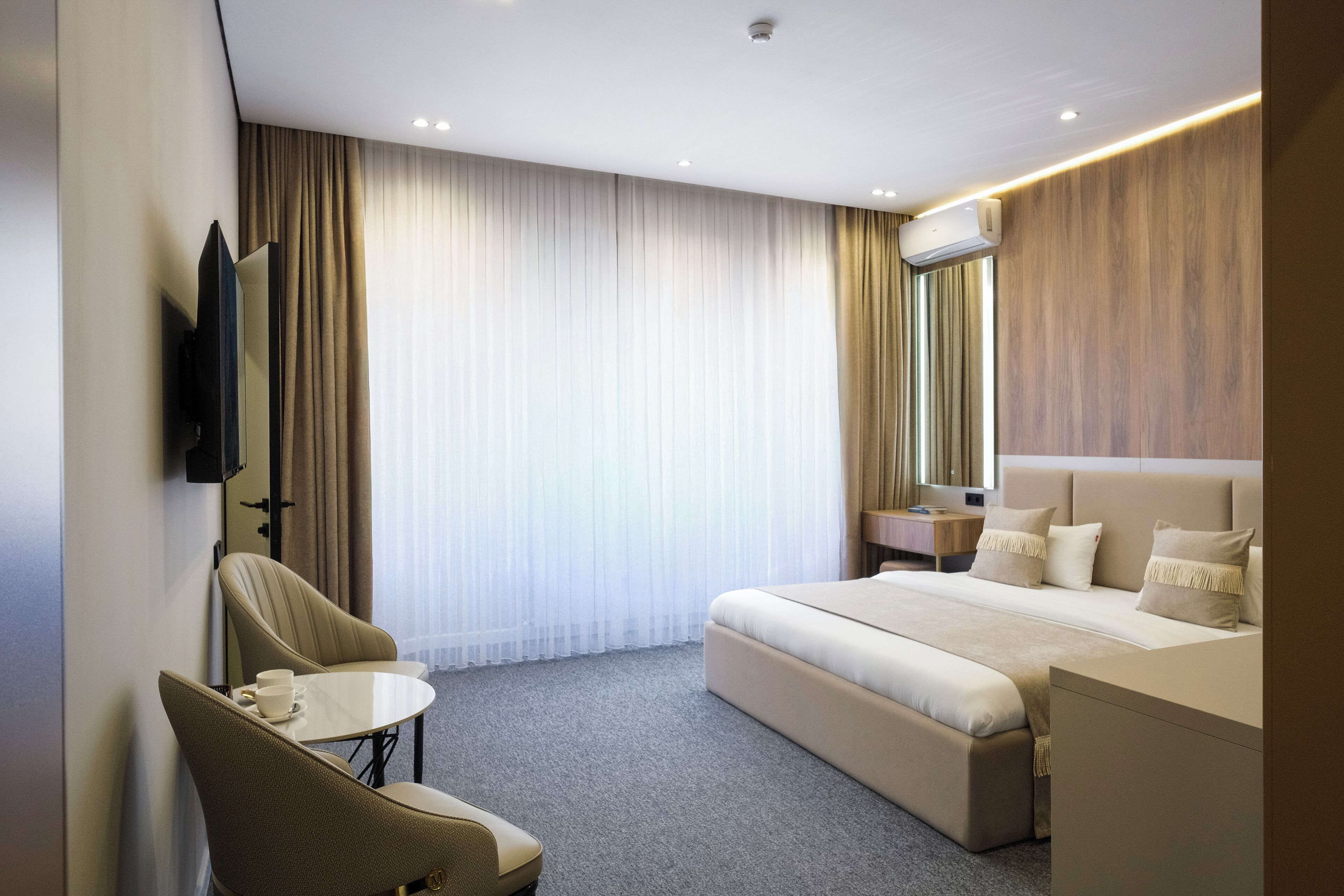A hotel room with a bed, a seating area with a table, a TV, and a window with curtains.