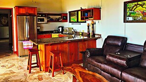 A kitchen with a refrigerator, microwave, and wooden cabinets. There is a bar area with stools and a leather couch. - Stunning Ocean View House with Pool, Outdoor Kitchen, and Rancho (Savegre de Aguirre)