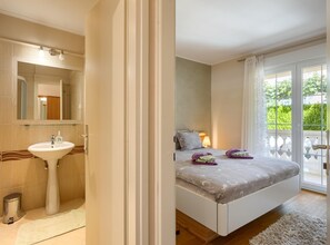 A bedroom with a bed, a nightstand, a lamp, and a view of greenery through the window. - Apartment in Porec Near Spadici Beach (Porec)