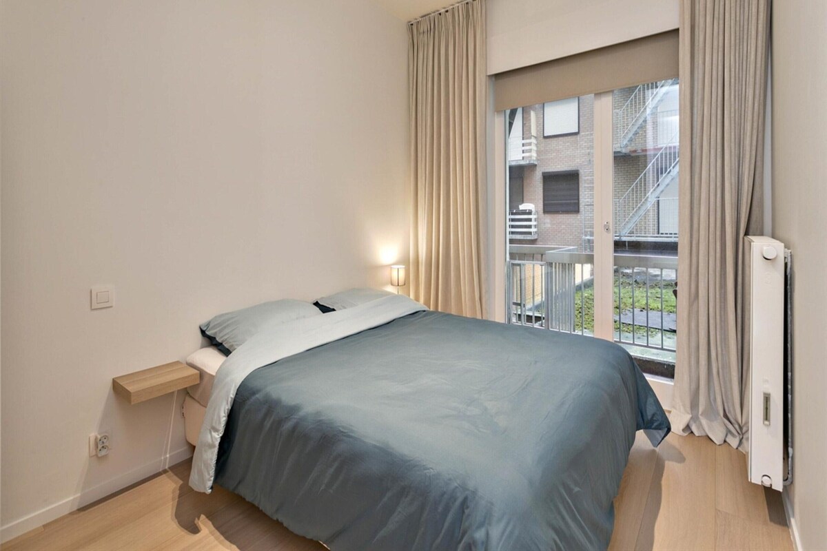 A bedroom with a bed, a bedside table, a window with curtains, and a radiator.