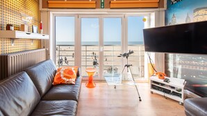 Una sala de estar con un sofá de cuero, una mesa de centro de vidrio, una televisión y un balcón con vista a la playa.