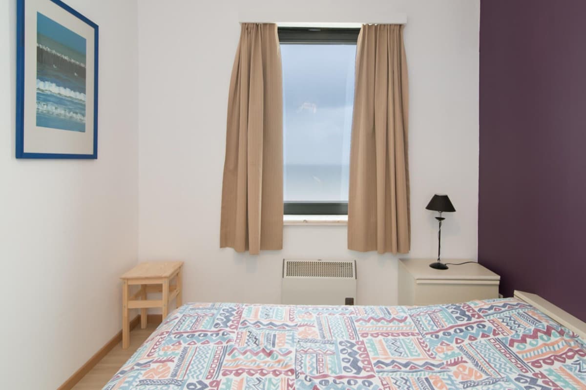 Ein Schlafzimmer mit einem Bett, einem kleinen Hocker, einem Nachttisch mit Lampe, einem Fenster mit Vorhängen und einer Heizung.