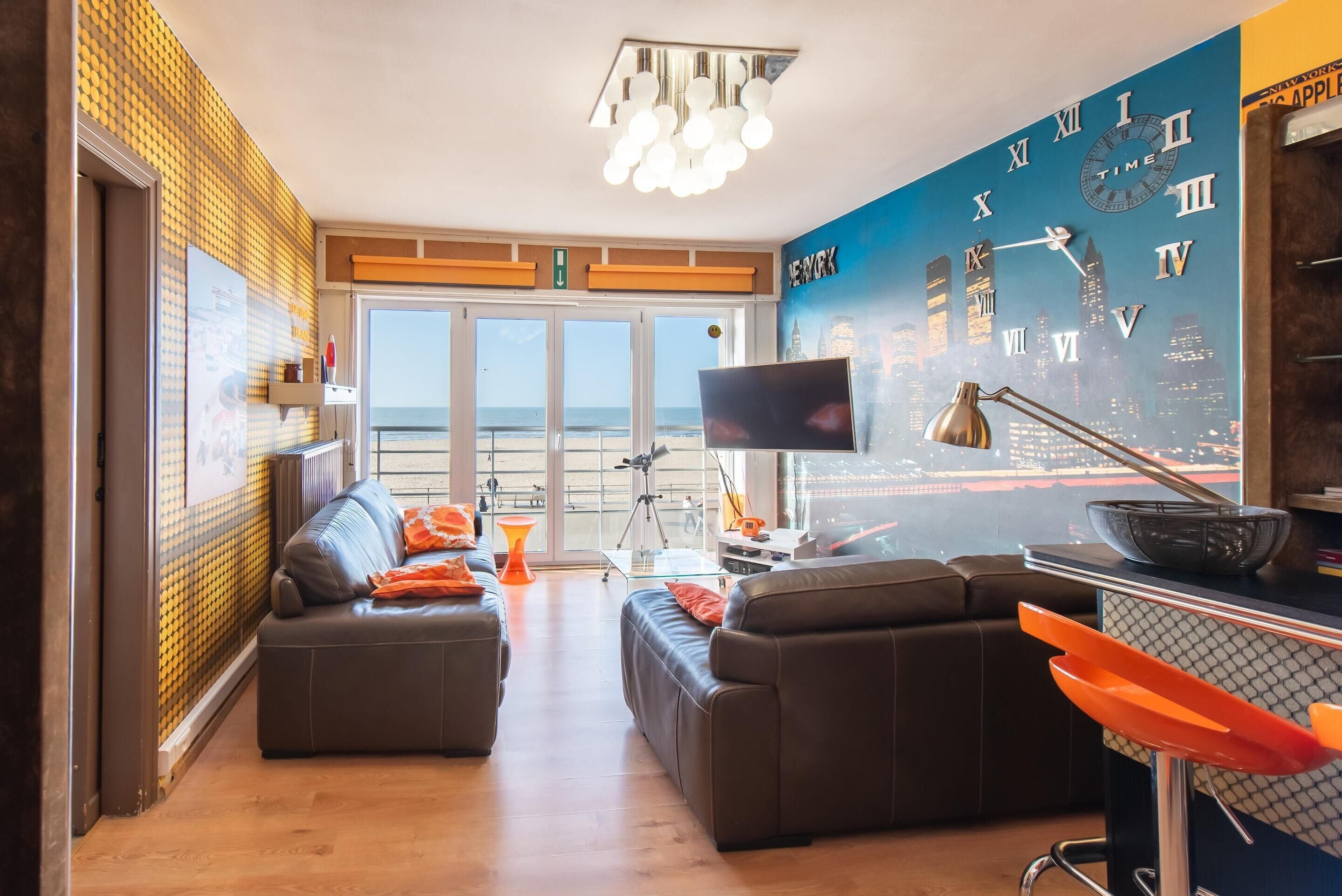Ein modernes Wohnzimmer mit Stadtlandschaftsmalerei, einem großen Flachbildfernseher und Blick auf den Strand durch große Fenster.