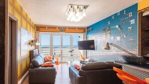 Un salon moderne avec une fresque représentant un paysage urbain, un grand téléviseur à écran plat et une vue sur la plage grâce à de grandes fenêtres.
