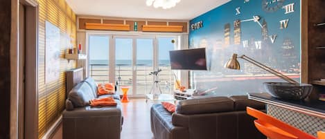 A modern living room with a cityscape mural, a large flat-screen TV, and a view of the beach through large windows.