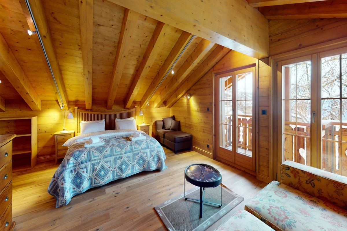 A wooden cabin bedroom with a bed, bedside table, lamp, and a view of the outside.
