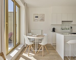 Eine moderne Küche mit weißen Schränken, ein Essbereich mit einem Holztisch und weißen Stühlen sowie ein Blick durch große Schiebeglastüren.
