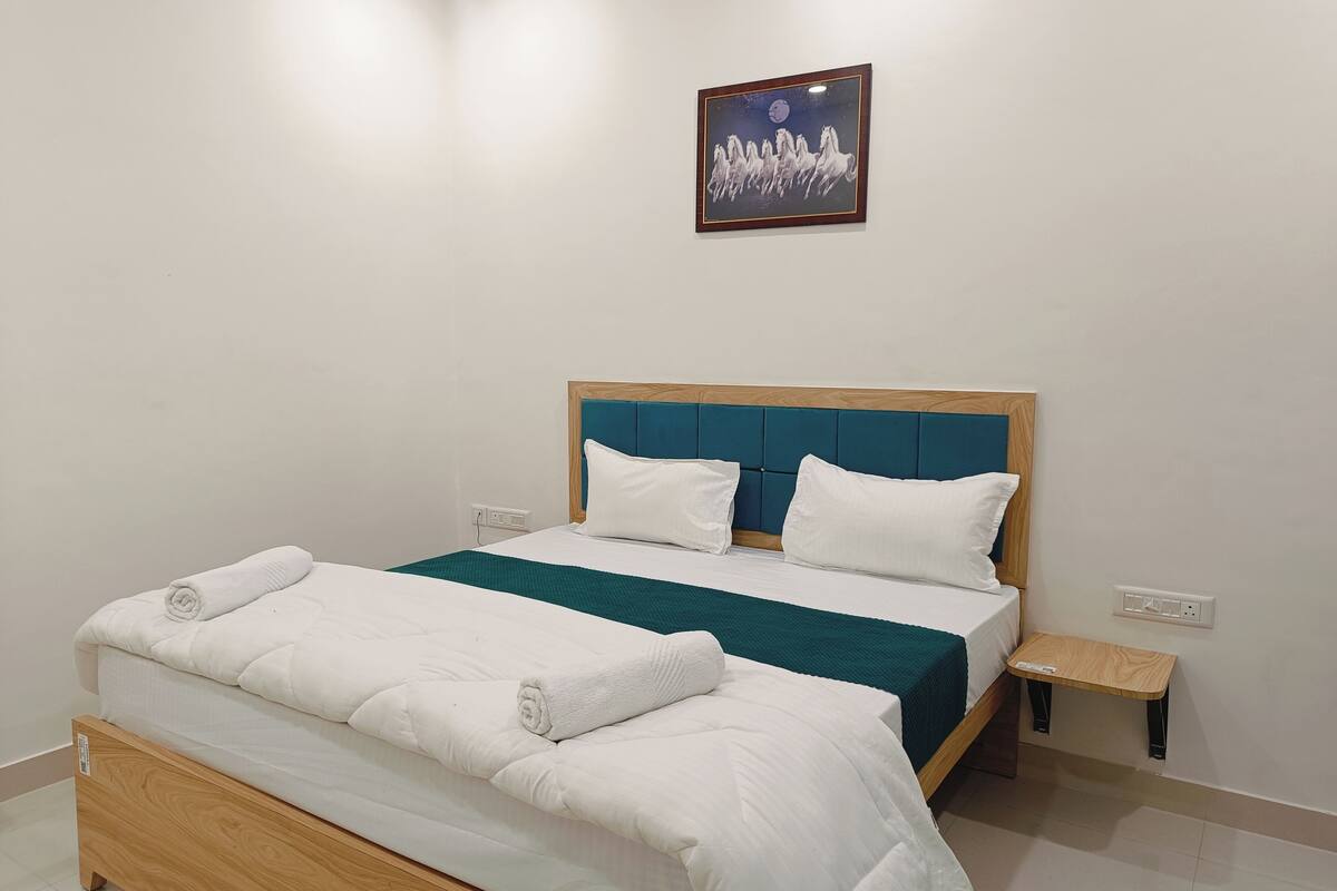 Een net opgemaakt bed met witte lakens en een blauwgroene plaid, een houten hoofdbord, een klein nachtkastje en een aan de muur hangend schilderij.