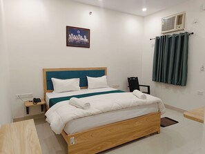 Ein ordentlich bezogenes Bett mit weißer Bettwäsche und einem Türkis-Akzent, ein Nachttisch mit Telefon und Ladegerät, eine wandmontierte Klimaanlage und ein gerahmtes Bild über dem Bett.