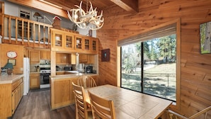 A wooden cabin interior with a dining area, kitchen, and a large window overlooking a forest. - Lakeview Haven- Luxury Retreat (Big Bear Lake)