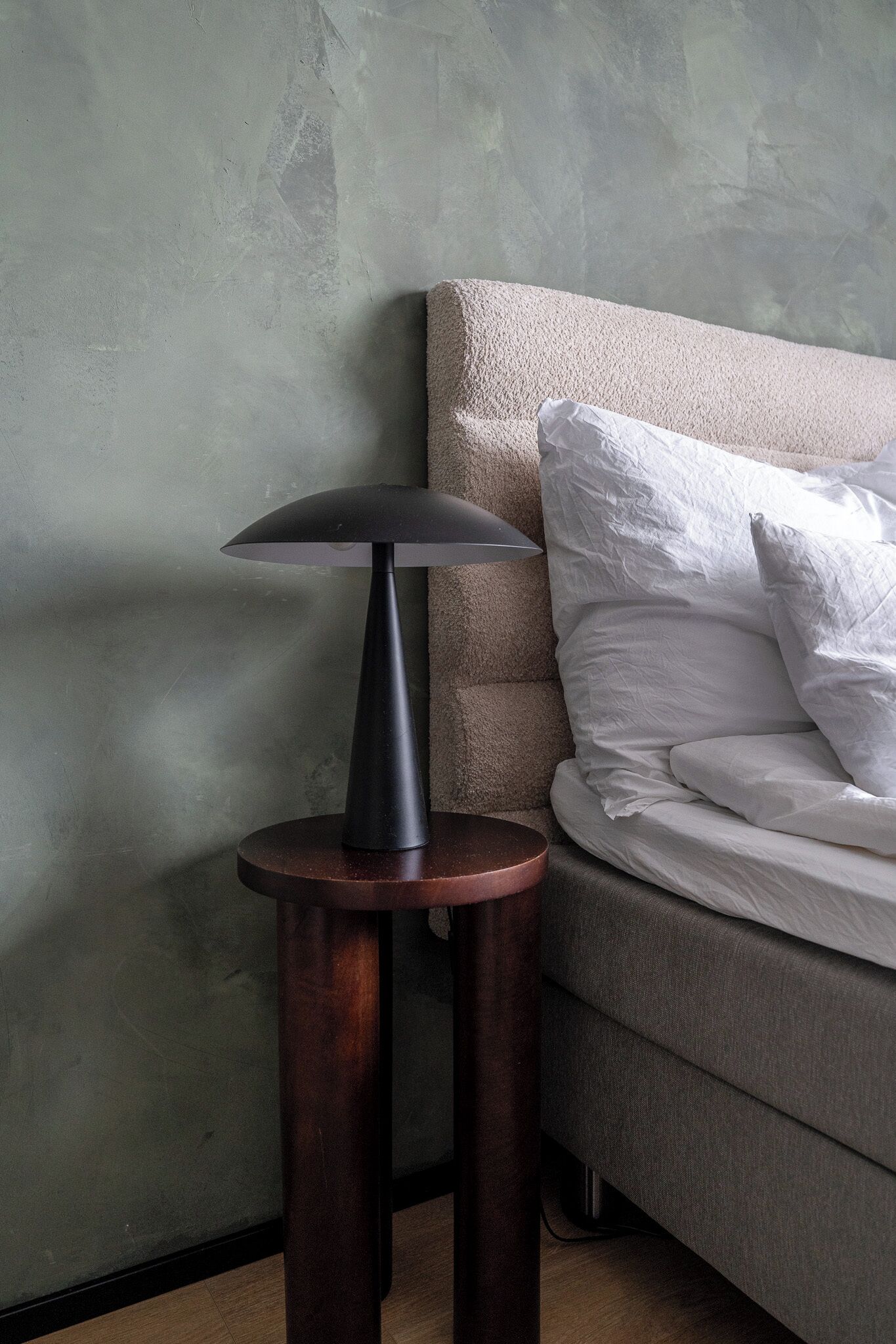 A modern lamp on a round wooden table beside a bed with a textured headboard.