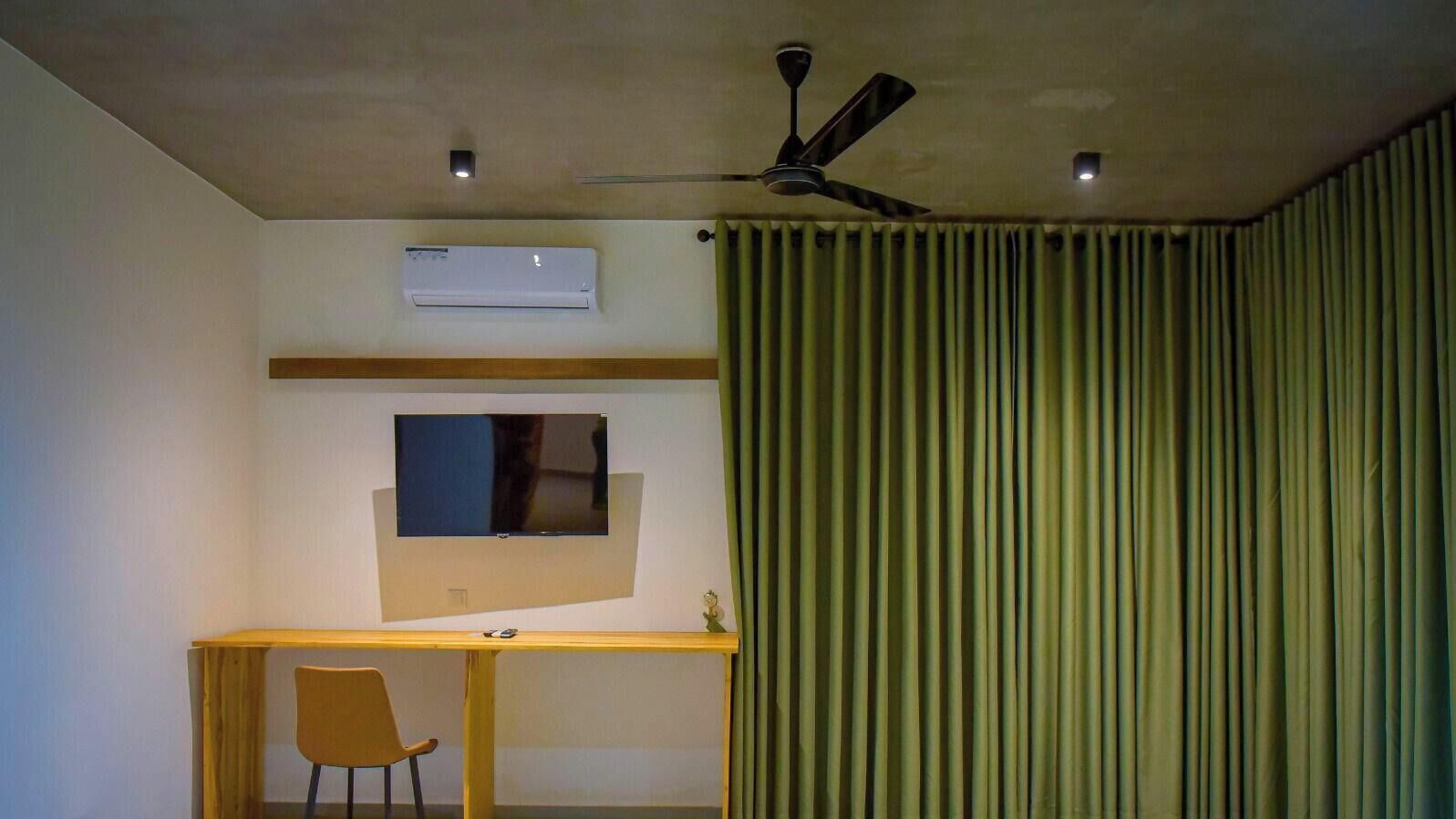 A room with a ceiling fan, an air conditioning unit, a flat-screen TV mounted on the wall, a desk with a chair, and a curtain.