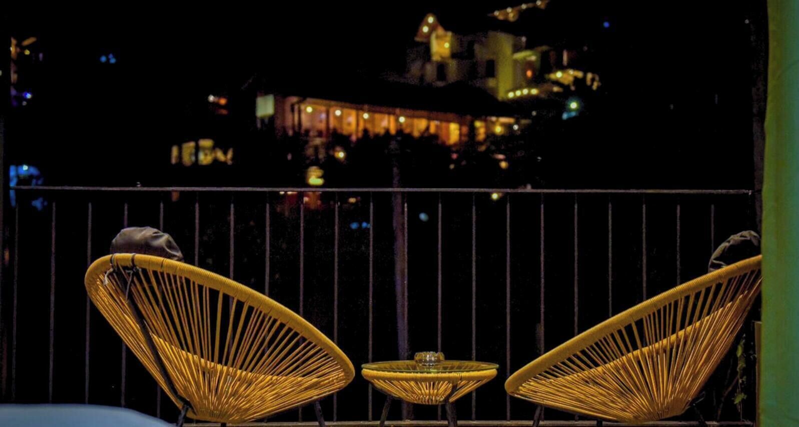 Un balcón con sillas de mimbre y una pequeña mesa, con vistas a un edificio iluminado por la noche.
