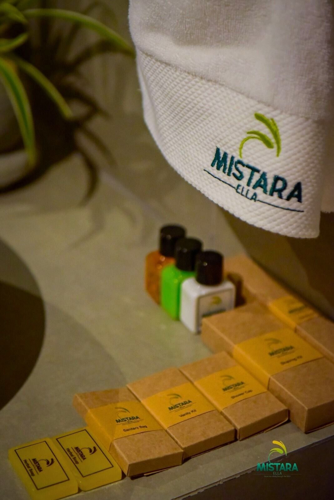 A towel with 'MISTARA' branding, a set of toiletries, and a plant in the background.