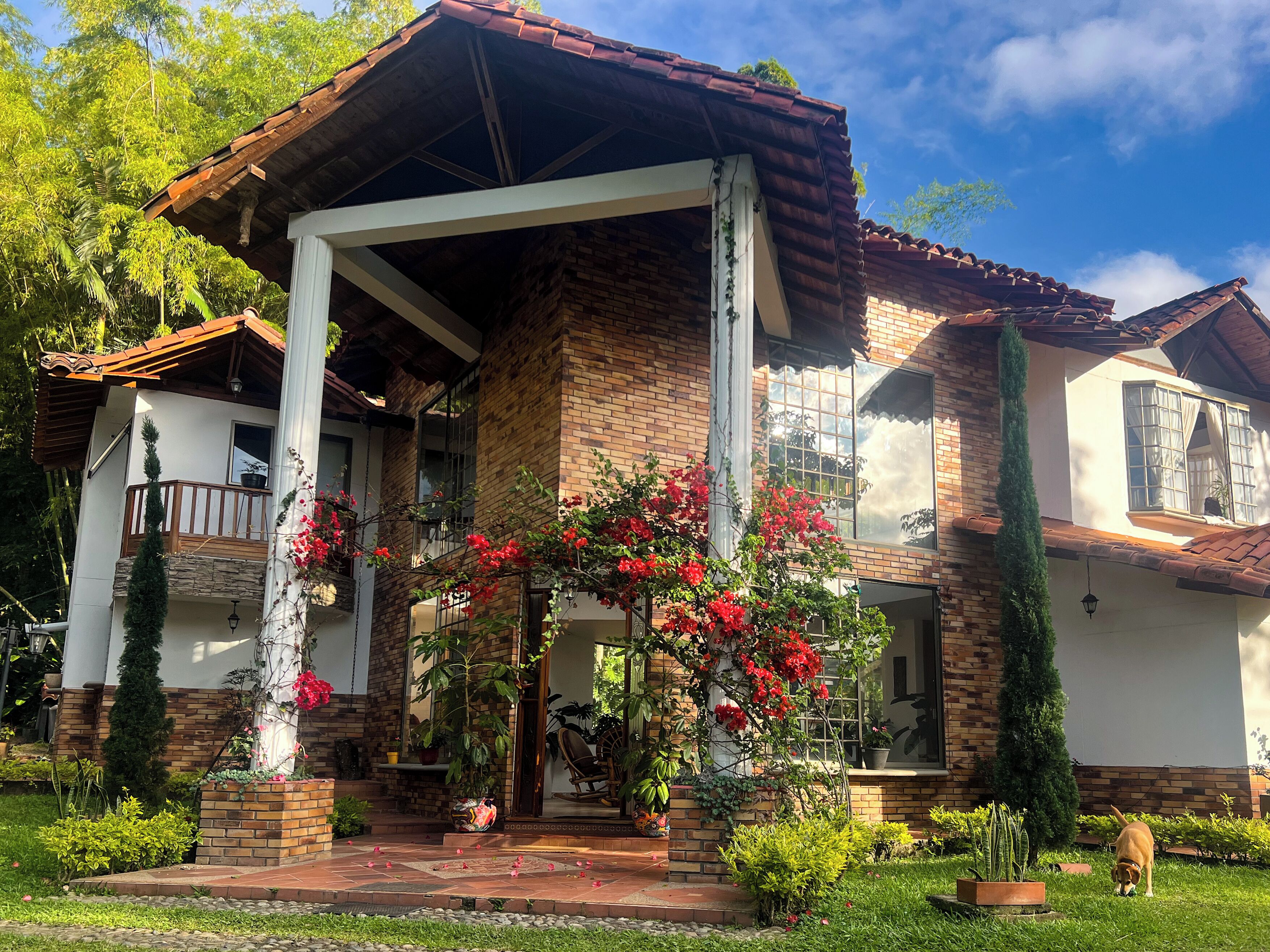 Une maison à deux étages avec une façade en briques, un balcon en bois et un jardin agrémenté de plantes en pot et de fleurs.