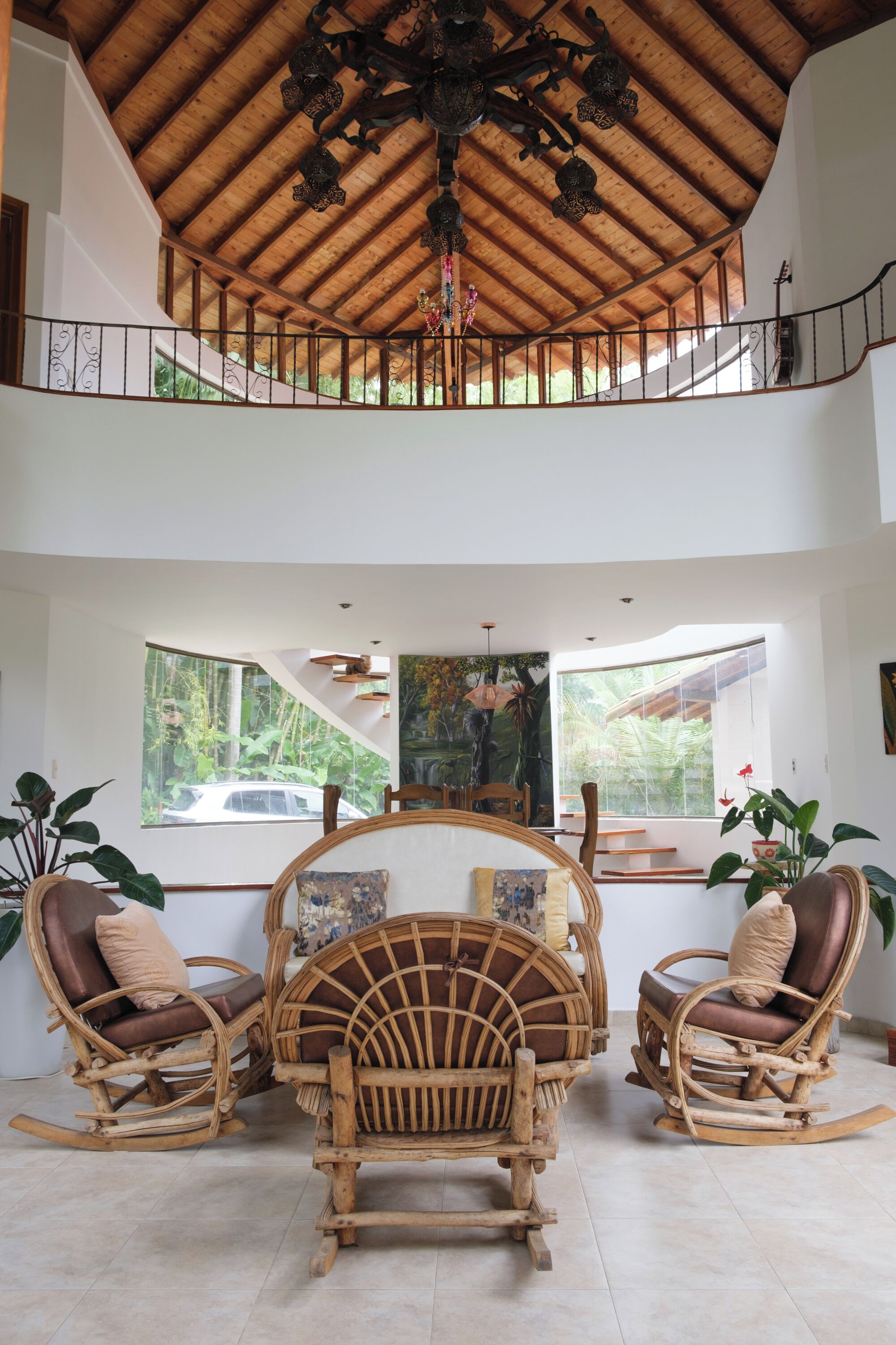 A spacious interior with a high wooden ceiling, a balcony, wicker furniture, and large windows offering views of the outdoors.