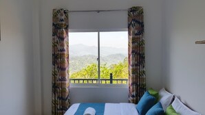 A bedroom with a bed, colorful curtains, and a view of greenery and mountains through a window.