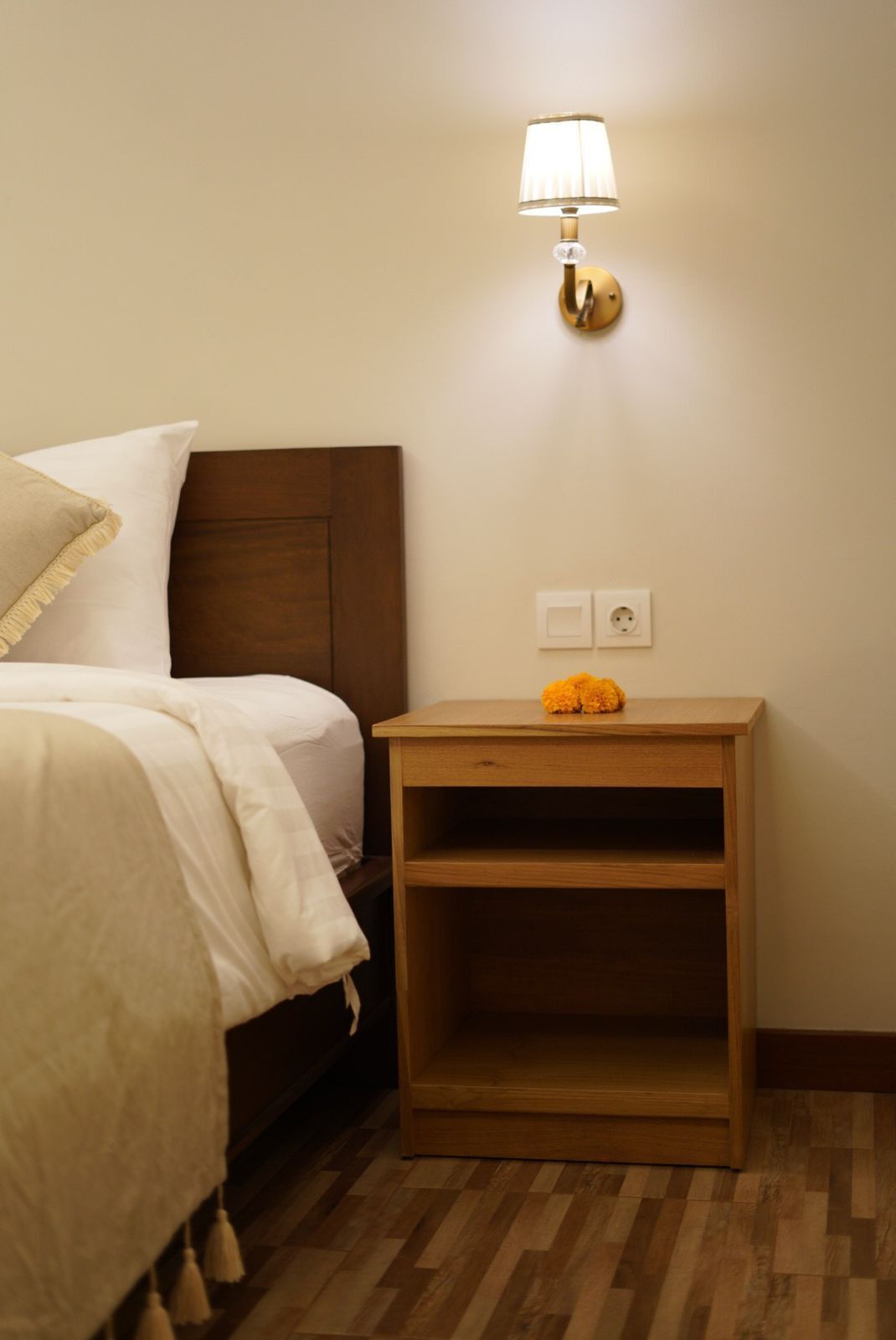 A neatly made bed with a headboard, a bedside table with a lamp, and a power outlet on the wall.