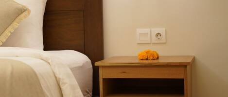 A neatly made bed with a headboard, a bedside table with a lamp, and a power outlet on the wall.