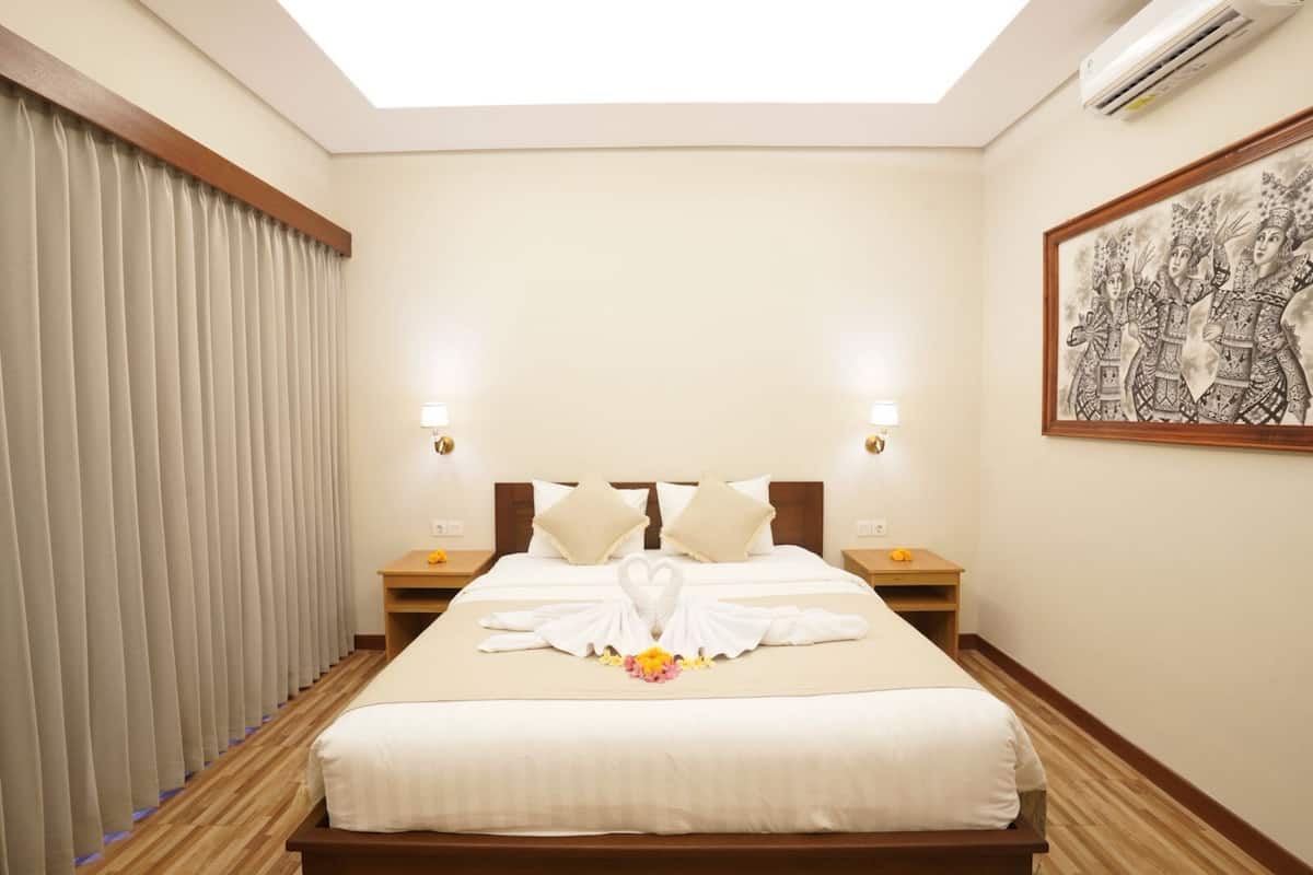 A hotel room with a large bed, two bedside tables, a ceiling light, an air conditioning unit, and a large window with curtains.