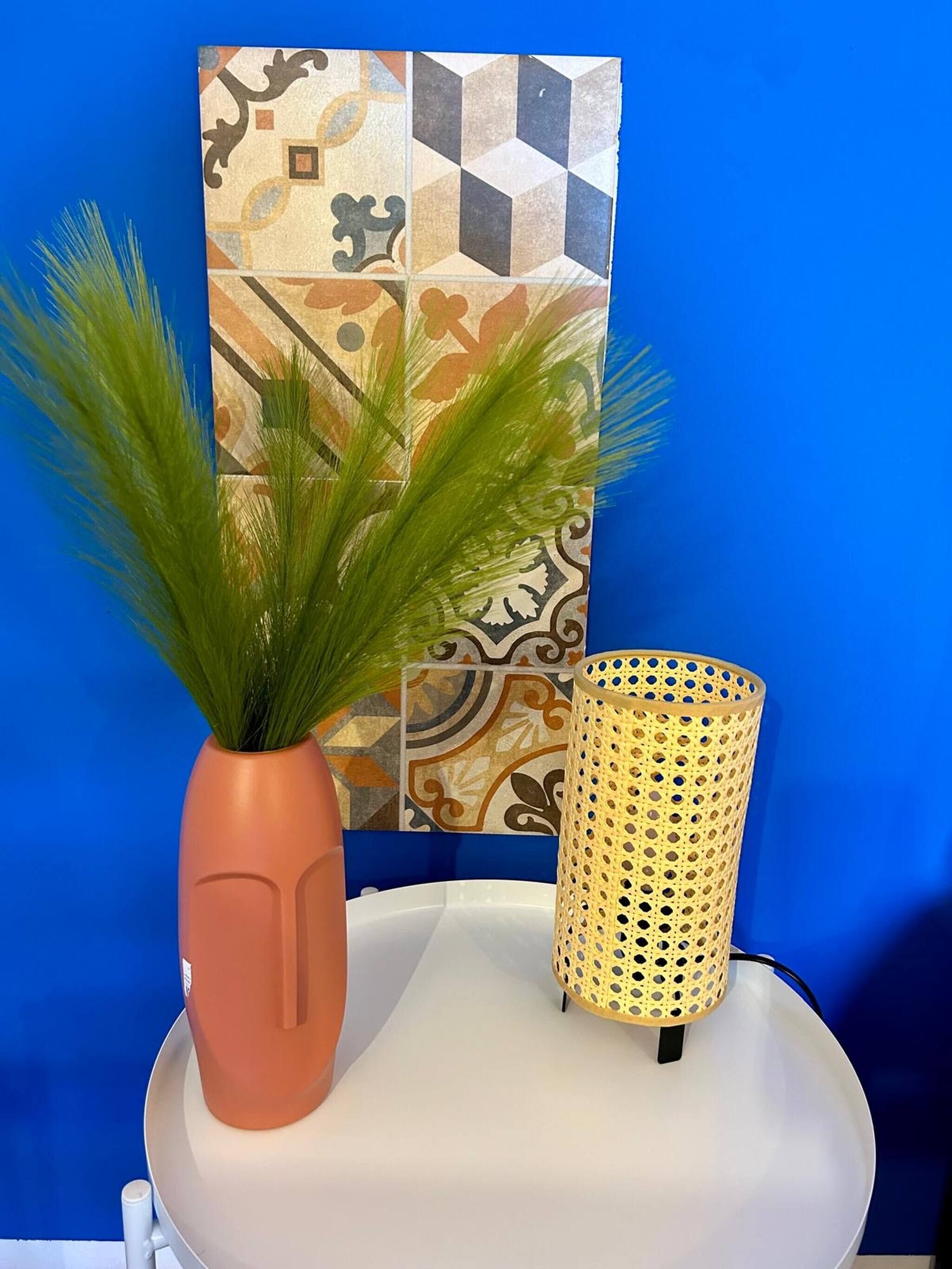 A white table with a potted plant and a perforated lamp against a blue wall with patterned tiles.
