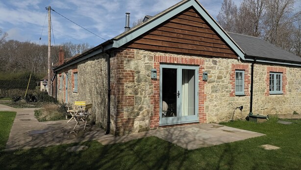Ein Steinhaus mit einem Holmgiebel, einer Tür mit Glasfenster und einer kleinen Außenbestuhlung.