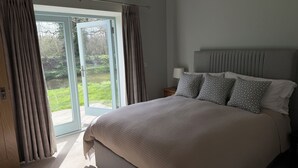 Ein Schlafzimmer mit einem großen Bett, einem Nachttisch, einer Lampe und Blick durch eine Glastür auf einen Garten.