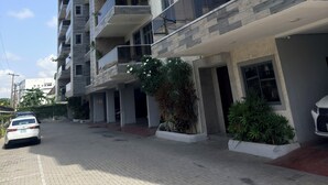 Un edificio residencial de varios pisos con balcones, un camino pavimentado y algo de vegetación.