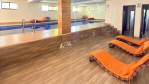 Una piscina interior con suelo de madera, tumbonas naranjas y una columna central.