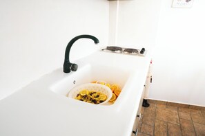 A kitchen sink with a colander containing food items, a black faucet, and a stove with two burners. - Traditional CycladicStudio-Zefyria (Zefiria Milos)