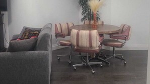 Ein Zimmer mit dunklem Holzboden, einem grauen Sessel, einem gestreiften Sofa, einem runden Tisch mit Stühlen, einer Topfpflanze und einem gerahmten Bild an der Wand.