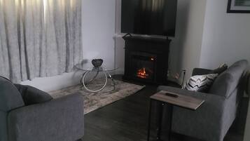 Un salon moderne avec une cheminée, un téléviseur à écran plat et deux fauteuils gris.