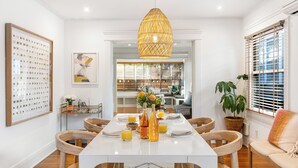 Een eetruimte met een witte tafel, houten stoelen, een hanglamp en uitzicht op de keuken.