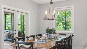Uma área de refeições com uma mesa de madeira, cadeiras, um lustre e uma janela com vista para árvores.