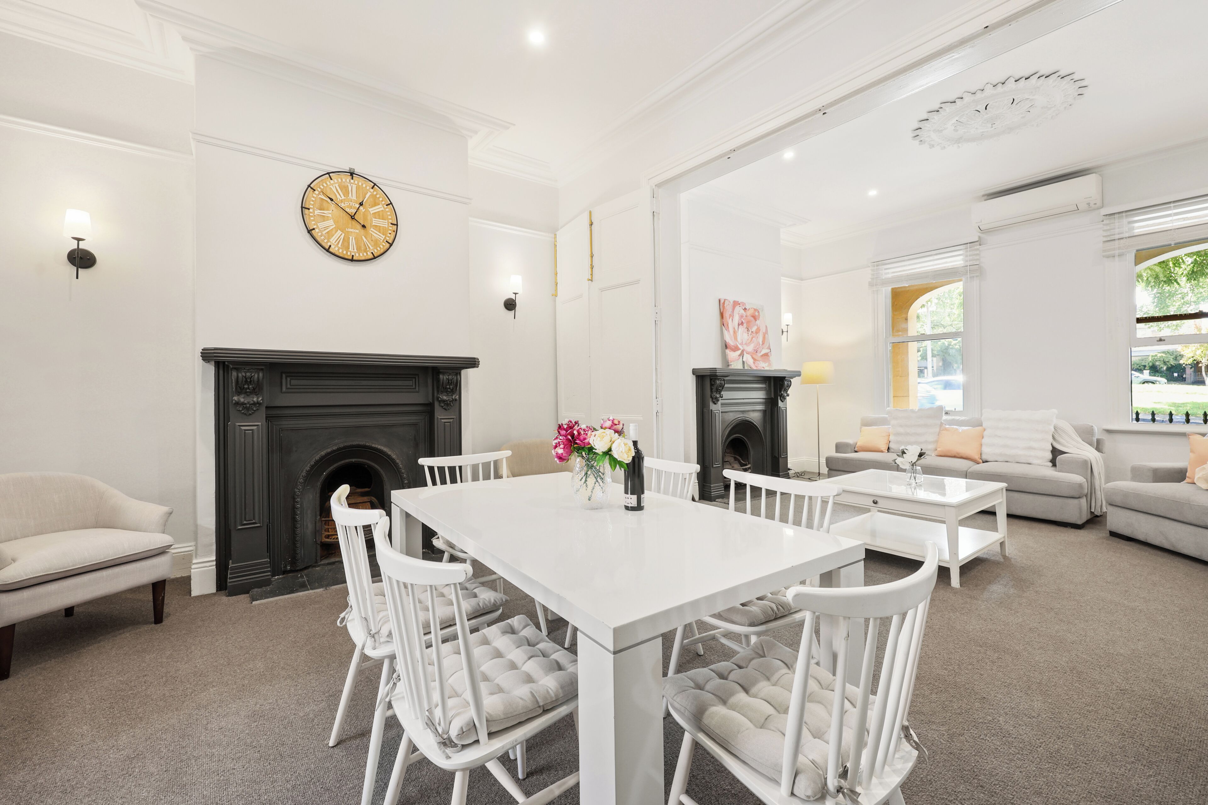 A spacious living room with a white dining table, white chairs, a fireplace, and a large clock on the wall.