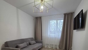 Ein modernes Wohnzimmer mit grauem Sofa, Holzboden, einer Deckenleuchte, einem Fenster mit Vorhängen und einem wandmontierten Fernseher.