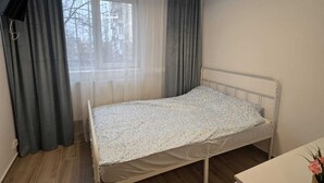 Ein Einzelbett mit einem gemusterten Überwurf, ein Nachttisch, ein Schreibtisch, eine Deckenleuchte und ein Fenster mit Vorhängen.