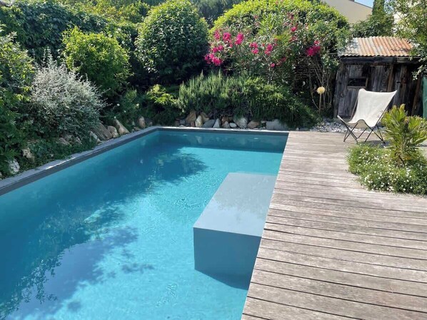A rectangular swimming pool with a wooden deck, surrounded by lush greenery and a small shed.