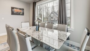 Comedor con una mesa de mármol, sillas tapizadas en gris, un cuadro de un pez y vistas a una casa a través de amplias ventanas.